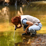 Frau mit rotem Haar beugt sich über einen Bach und misst das Wasser mit einem Handgerät. Herbstblätter um sie herum.