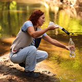 Frau füllt eine Wasserflasche an einem ruhigen Gewässer mit einem Filter. Die Umgebung ist grün und naturbelassen.