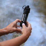 Person montiert einen BOXIO Wasserfilter auf eine transparente Wasserflasche am Ufer eines Gewässers.